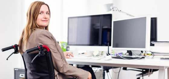 Inhaltsbild: Eine Frau im Rollstuhl sitzt am Schreibtisch vor dem Computer und lächelt in die Kamera.