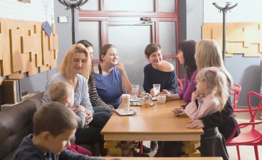 Thumbnail: Mamas sitzen mit ihren kleinen Kindern am Tisch und unterhalten sich bei einem Kaffee.