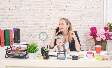 Thumbnail: Eine Mutter sitzt am Schreibtisch und hat ihr kleines Baby auf dem Schoß, einen Stift in der einen und das Telefon in der anderen Hand.
