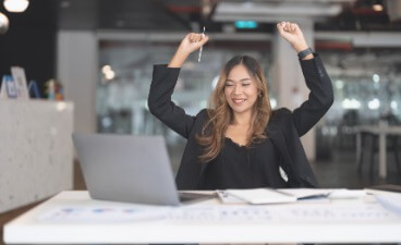 Thumbnail: Eine Frau mit schwarzem Blazer sitzt am Schreibtisch vor dem Laptop im Büro und reißt lächelnd die Hände in die Luft.