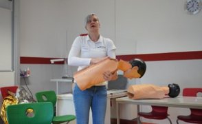 Eine Frau hält eine Übungspuppe im Arm und erklärt die erste Hilfe bei Verschlucken eines Fremdkörpers