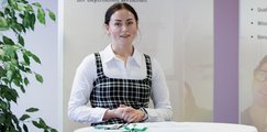 Auf diesem Bild ist eine junge Frau an einem Stehtisch in einem Büro zu sehen. Sie trägt ein kariertes Kleid und schaut in die Kamera. Hinter ihr ist ein Informationsplakat sichtbar, das den Schriftzug "Die Beruflichen Fortbildungszentren der Bayerischen Wirtschaft" trägt.