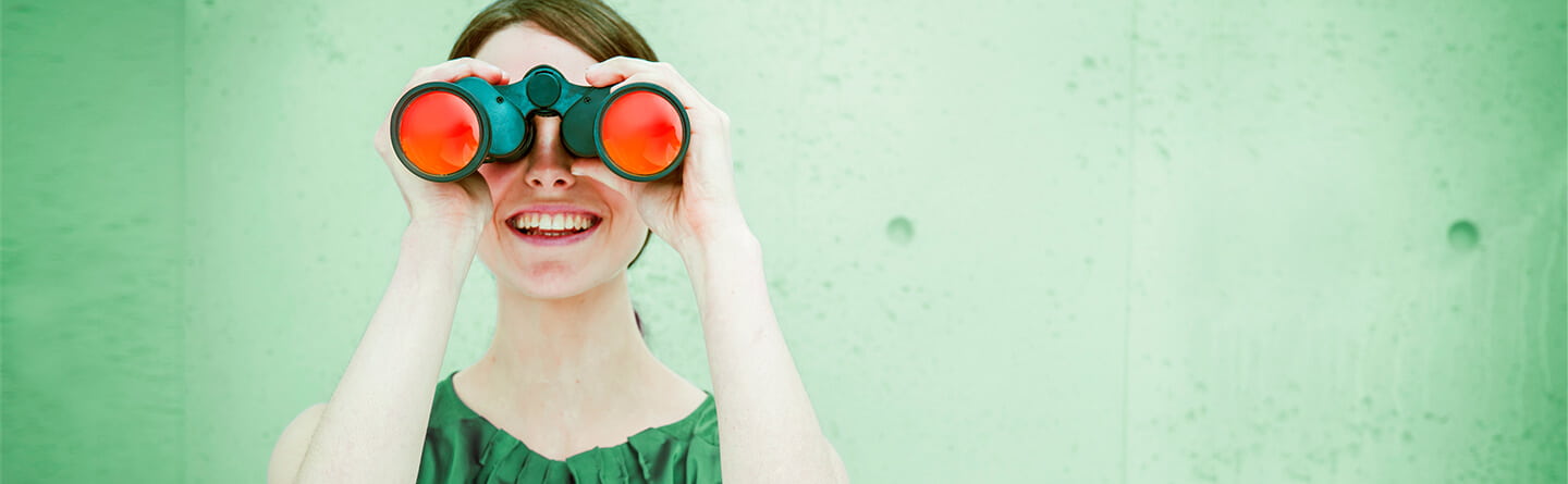 Header: Lächelnde Frau vor grünem Hintergrund hält grünes Fernglas mit roten Gläsern in Händen.