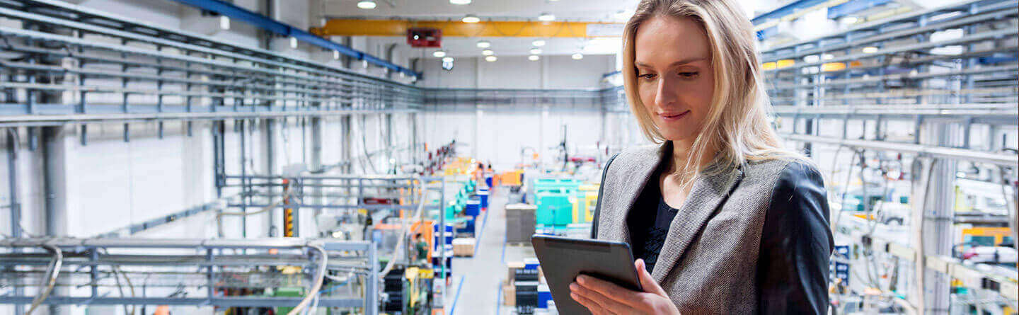 Header: Frau in einer Fabrikhalle mit Maschinen hält ein Tablett um die Produktion zu Überprüfen. 
