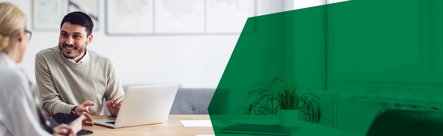 Grundsicherung Header: Ein Mann am Laptop spricht mit einer Frau in einem Büro