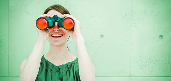 Inhaltsbild: Lächelnde Frau vor grünem Hintergrund hält grünes Fernglas mit roten Gläsern in Händen.