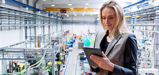 Inhaltsbild: Frau in Fabrikhalle mit Maschinen hält ein Tablett um die Produktion zu Überprüfen.