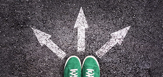 Inhaltsbild: Grüne Schuhe mit 3 in verschiedene Richtungen zeigende Pfeile auf dem Asphaltboden.