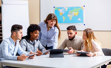 Eine Schülergruppe sitzt an einem Tisch und arbeitet mit Tablets, während eine Lehrkraft daneben steht und unterstützt.
