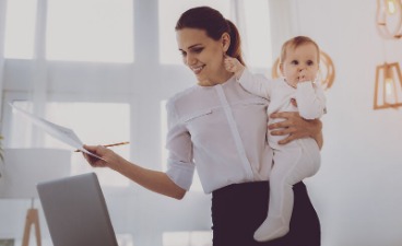 Eine Frau hält ein Baby auf dem Arm und arbeitet gleichzeitig mit Unterlagen an einem Laptop.