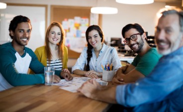 Fünf Personen sitzen gemeinsam am Tisch, lächeln in die Kamera und arbeiten entspannt an Ideen und Unterlagen.