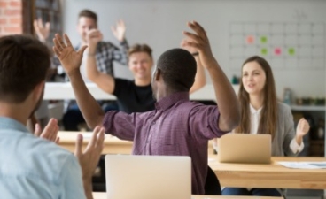 Teilnehmende in einem Workshop sitzen an Tischen und melden sich aktiv mit erhobenen Händen.