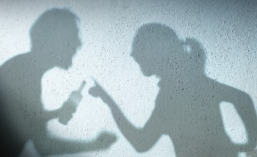 Die Schatten zweier Personen streiten heftig; eine Silhouette hält eine Flasche, die andere zeigt anklagend mit dem Finger.