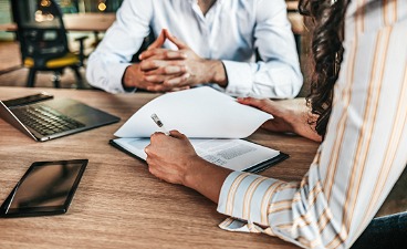 Zwei Personen sitzen an einem Tisch, eine schreibt in ein Dokument, Laptop und Smartphone liegen daneben.
