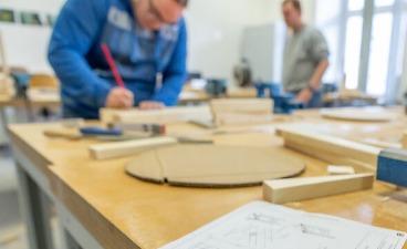 Eine Person arbeitet in einer Werkstatt an Holzbauteilen, im Vordergrund liegen Werkstücke und technische Zeichnungen.