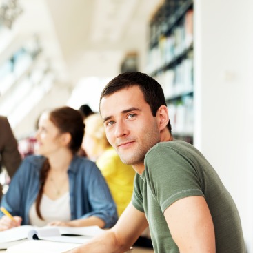Ein junger Mann schaut in die Kamera, im Hintergrund erklärt eine Dozentin oder Lehrerin einer jungen Frau etwas.