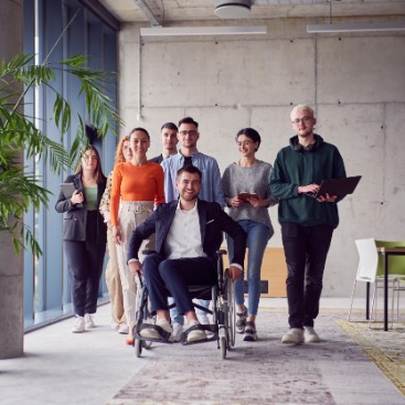 Eine diverse Gruppe steht in einem Seminarraum, darunter auch eine im Rollstuhl sitzende Person.