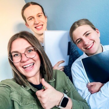 Drei Schüler*innen lachen in die Kamera, zwei von ihnen halten Hefter in den Händen, ein Mädchen hält den Daumen nach oben.