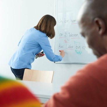 Lernsituation Deutschkurs, Frau schreibt was an die Tafel. Mann hört zu.