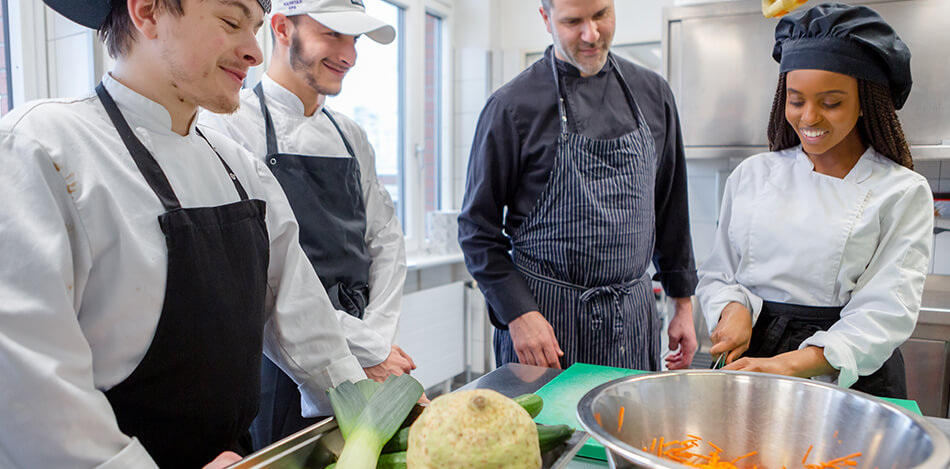 Koch erklärt Auszubildenden die Technik des Gemüseschneidens.