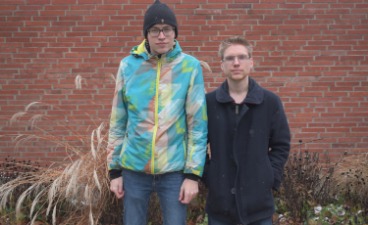 Zwei junge Männer stehen vor einer Backsteinwand.