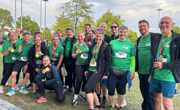 Gruppe von Teilnehmenden in grünen Laufshirts mit Medaillen beim Firmenlauf, posierend im Freien neben einer Laufstrecke