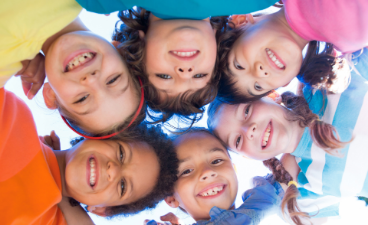 Sechs Kinder stehen im Freien in einem Kreis und blicken von oben in die Kamera vor blauem Himmel.