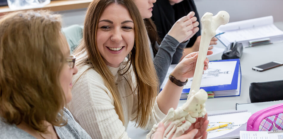 Schülerinnen unterhalten sich und lachen während eine der beiden im Anatomie-Unterricht einen künstlichen Knochen des Lehr-Skeletts in der Hand hält..