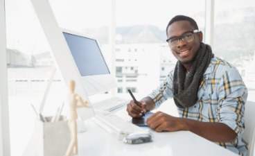 Teaserbild: Junger Mann sitzt in einem modernen Büro an einem Computer und lächelt in die Kamera. 