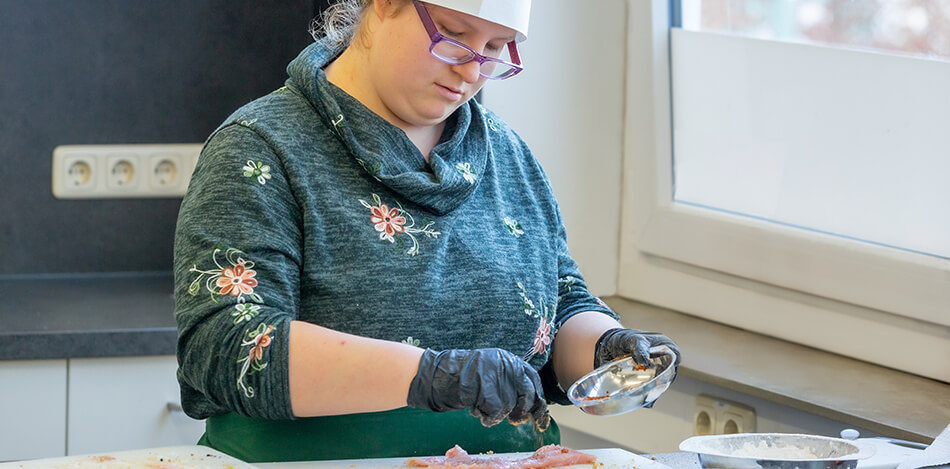 Bildgalerie: Junge Frau in einer Küche mit Kochmütze und Kochschürze würzt Fleisch.