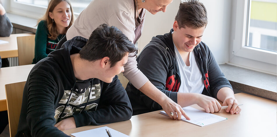 Bildergalerie: In einem Klassenzimmer zeigt eine Lehrerin zwei Schülern etwas auf einem Papier.
