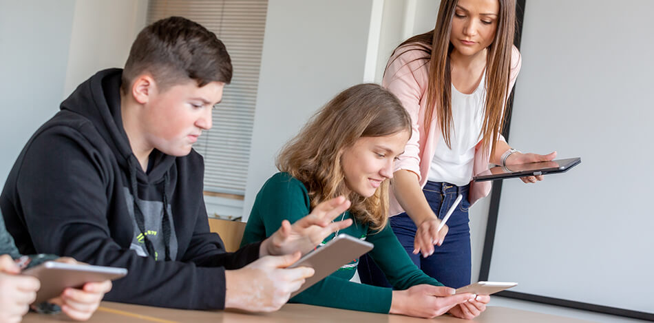 Bildergalerie: Eine Lehrerin erklärt zwei Jugendlichen in einem Klassenzimmer etwas auf dem Tablet.