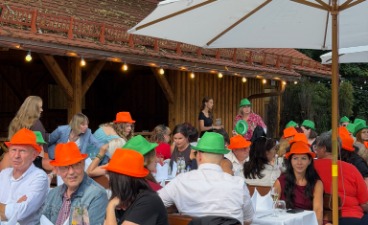 Mitarbeiter*innen sitzen am weiß gedeckten Tischen im Biergarten mit orangen und grünen Sommerhüten