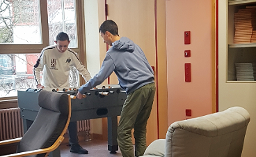 News- Foto: Zwei junge Männer spielen Kicker vor einer Fensterfront