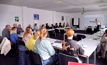 News- Foto: Teilnehmende sitzen an U-förmig angeordneten Tischen in einem Schulungsraum und verfolgen eine Präsentation, die an die Wand projiziert wird
