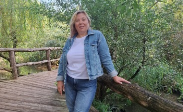 Eine Frau mit Jeansjacke und blonden Haaren steht vor Bäumen und lächelt in die Kamera.