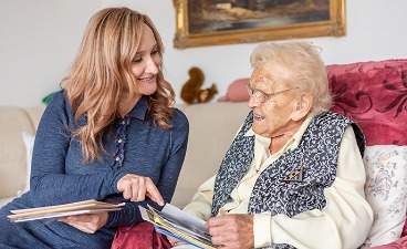 Eine Pflegekraft liest einer Seniorin aus einem Buch vor.