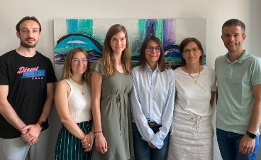 Vier Frauen und zwei Männer stehen vor einer Wand und lächeln in die Kamera. 