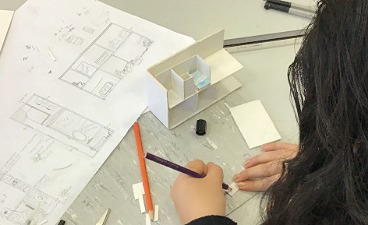 Teaserbild: Arbeitsplatz mit Bauplan und Schülerin, die ein kleines Haus nachbaut