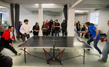 News- Foto: Um eine Tischtennisplatte, an der ein Doppel gespielt wird, stehen viele Jugendliche und feuern die Spielenden an