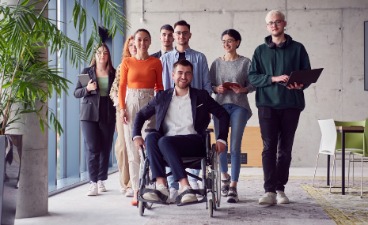 Eine diverse Gruppe steht in einem Seminarraum, darunter auch eine im Rollstuhl sitzende Person.