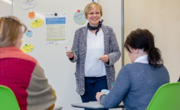 Eine Frau steht vor einem Whiteboard und spricht zu mehreren Personen.