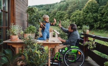 Teaser: Ein Mann im Rollstuhl und ein Kind geben sich auf einer Terrasse ein High Five.