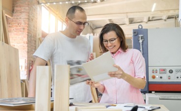 Zwei Personen betrachten gemeinsam Unterlagen in einer Werkstatt. Im Hintergrund sind Maschinen und Arbeitsmaterialien zu sehen.
