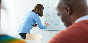Thumbnail: Eine Dozentin steht an einem Whiteboard und im Vordergrund sitzt ein Mann.