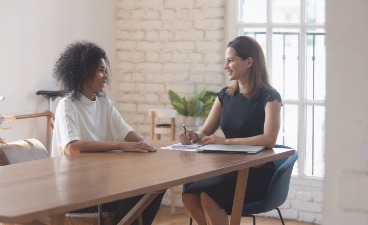 Thumbnail: Zwei Frauen sitzen an einem Tisch und unterhalten sich.