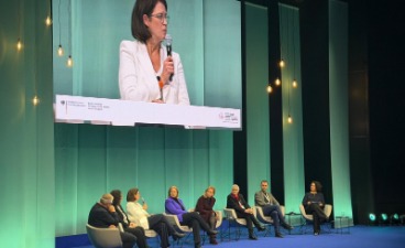 Eine Gruppe aus Männern und Frauen sitzen in einem Halbkreis auf einer Bühne und tauschen sich aus.