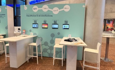 Ein Messestand aus weißen Stühlen und Tischen, mit einer hellblauen Messewand mit Tablets.