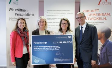 Das Bild zeigt vier Personen, die sich vor einem Banner mit den Worten "Wir eröffnen Perspektiven" und "Zukunft. Gemeinsam. Entwickeln." aufgestellt haben. Zwei Frauen und zwei Männer halten ein großes Förderbescheidscheck, der eine Summe von 1.199.168,97 € anzeigt. Die Person auf der linken Seite trägt ein rotes Blazer und die zweite eine graue Bluse, während die beiden Männer in formellen Anzügen gekleidet sind.