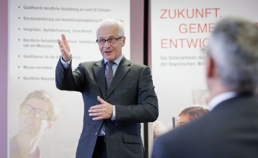Einblicke in den Arbeitsalltag der Anerkennungsberatung Das Bild zeigt einen Mann in einem grauen Anzug, der gestikulierend spricht. Er steht vor einem Plakat mit dem Schriftzug „Zukunft. Gemeinsam. Entwickeln.“ und scheint eine Diskussion oder Präsentation zu halten. Im Vordergrund ist die Person eines Zuhörers zu sehen, deren Gesicht nur teilweise sichtbar ist.